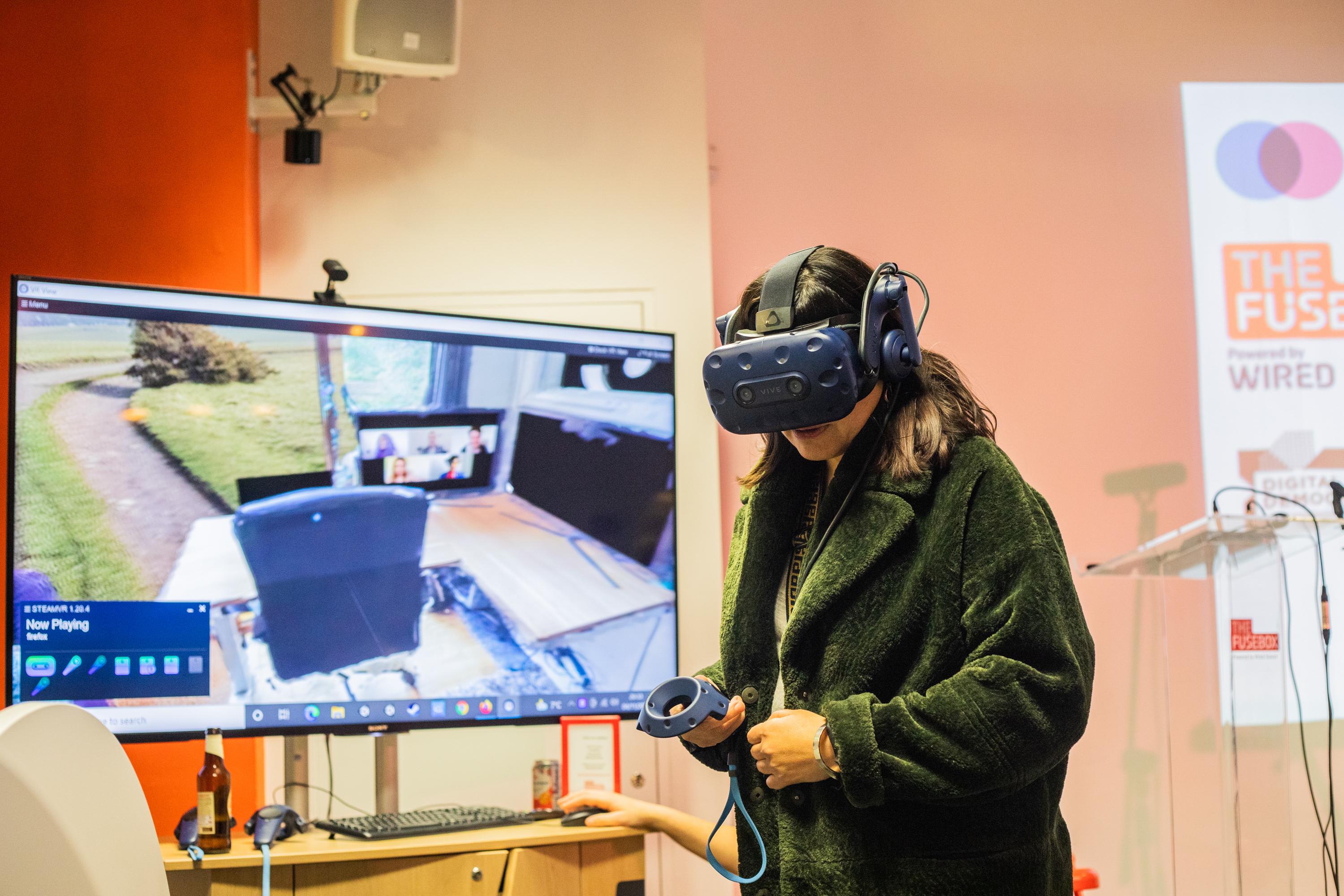 Woman wearing green jacket and VR headset standing in front of a screen where you can see what the person is experiencing in the Metaverse