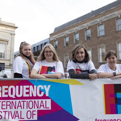 four young women wearing Frequency t-shirts, leaning on a Frequency Festival banner