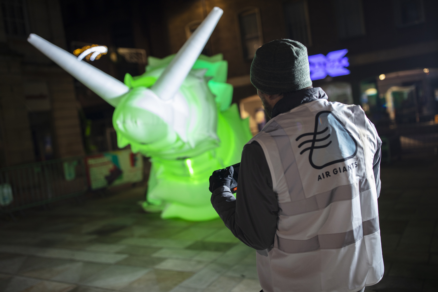 Foreground: Man standing with his back to the camera holding a control pad. Background: A giant inflatable robot snail lit up with a green light.
