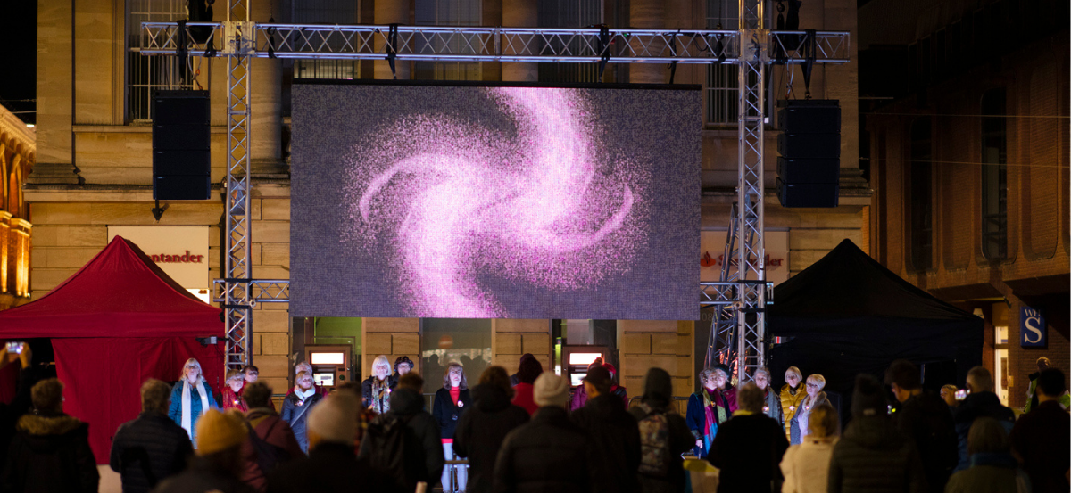 WE-Hope performance with a digital screen and choir outside in a public space in front of a building. Screen shows a swirling pink visual graphic.