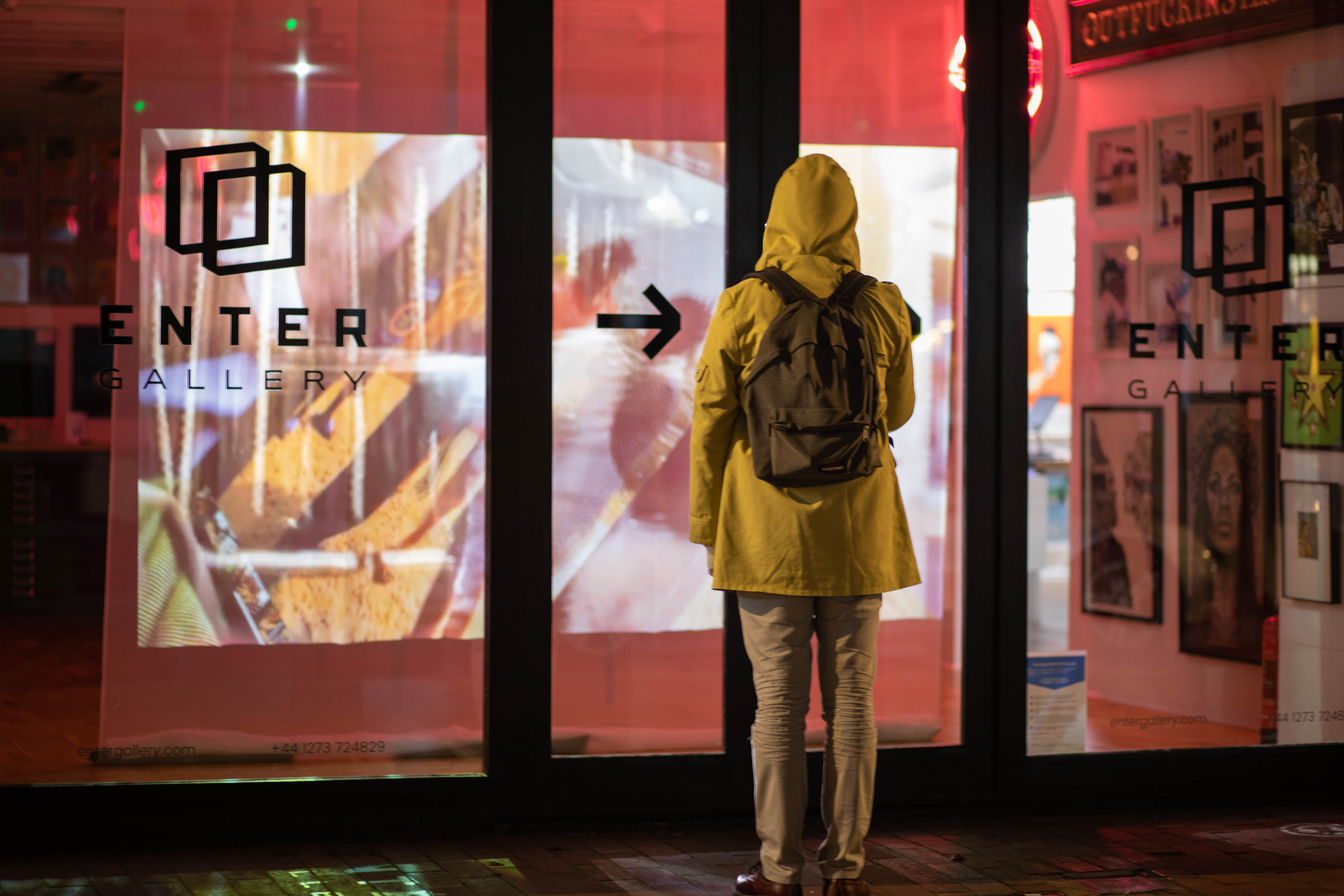 person wearing yellow coat and a back pack, standing in front of a gallery window showing a projected screen behind the glass. Text on window says 'Enter Gallery' with an arrow pointing to the right.