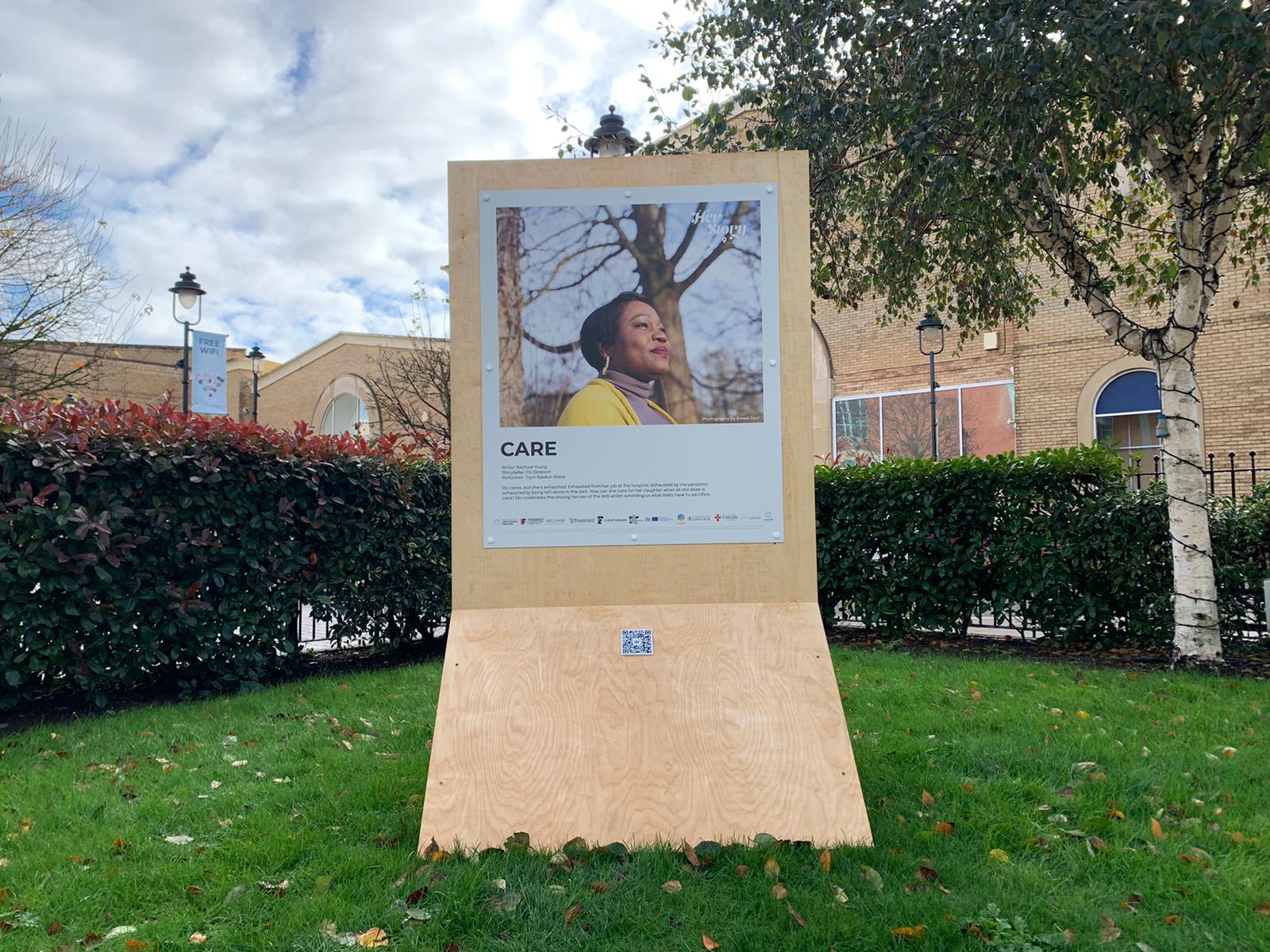 wooden board with photo of a woman and text beneath, in middle of a park.