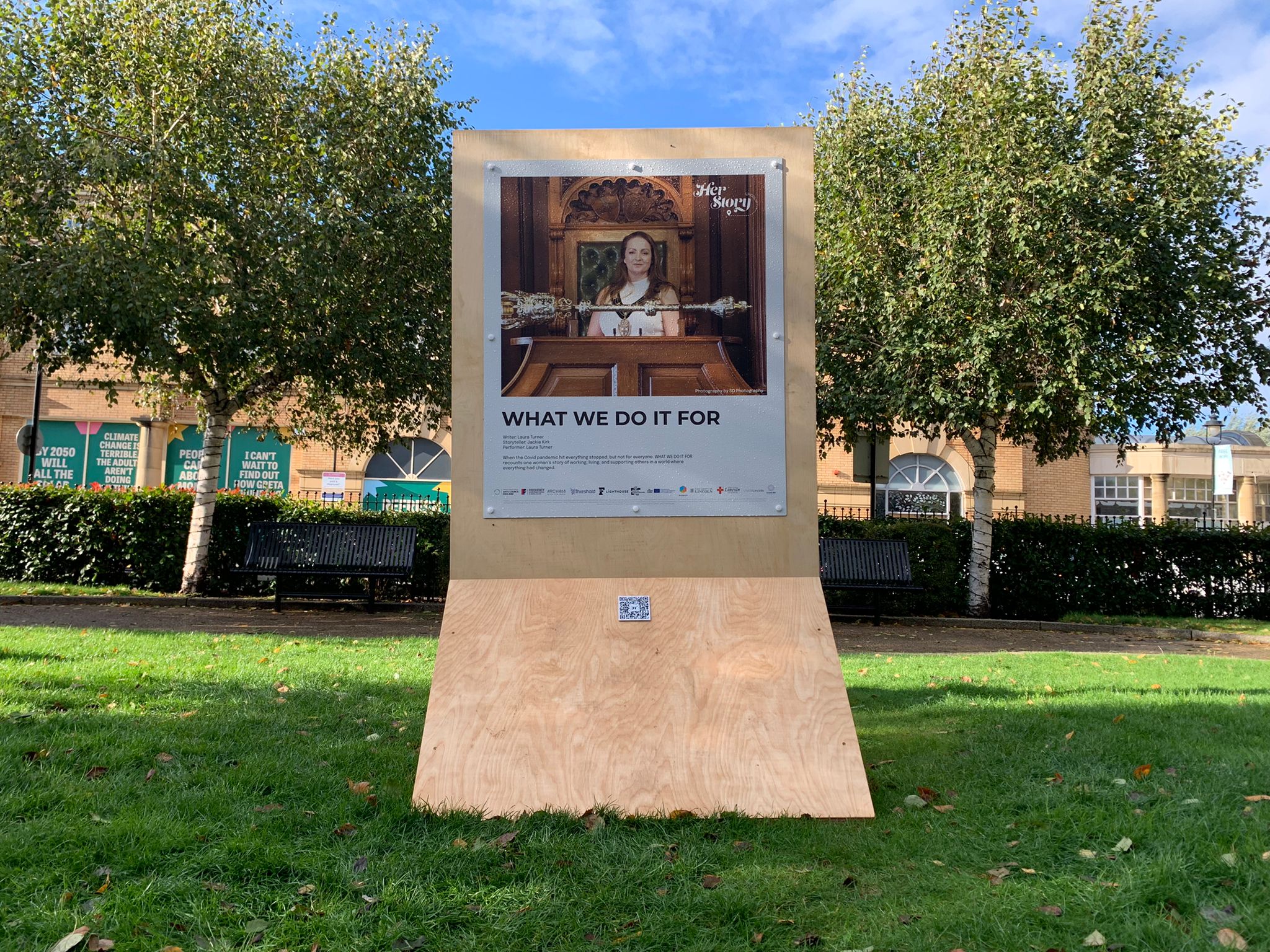 wooden board with photo of a woman and text beneath, in middle of a park.