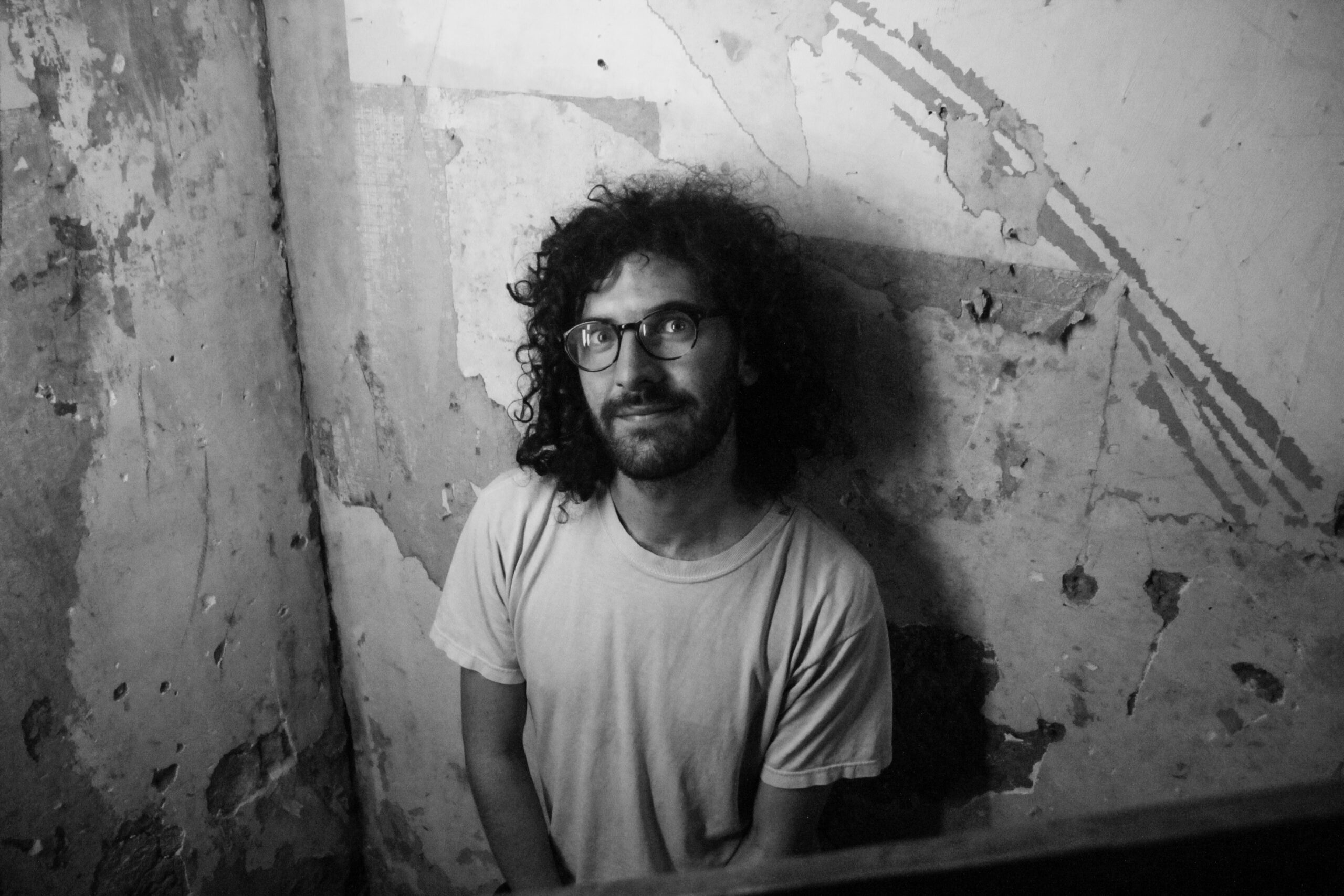 black and white photo of a person with long curly hair, a beard and glasses wearing a white t-shirt.