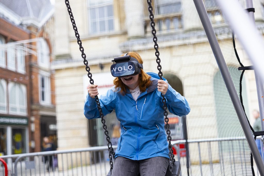 Person with long red hair and a blue rain jacket on a swing in Cornhill Quarter Lincoln, wearing a VR headset
